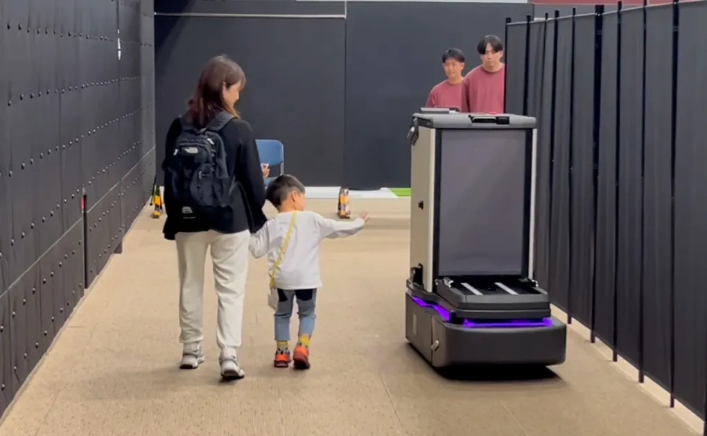 廊下を歩いている親子と走行中のモノを運ぶロボットの写真。